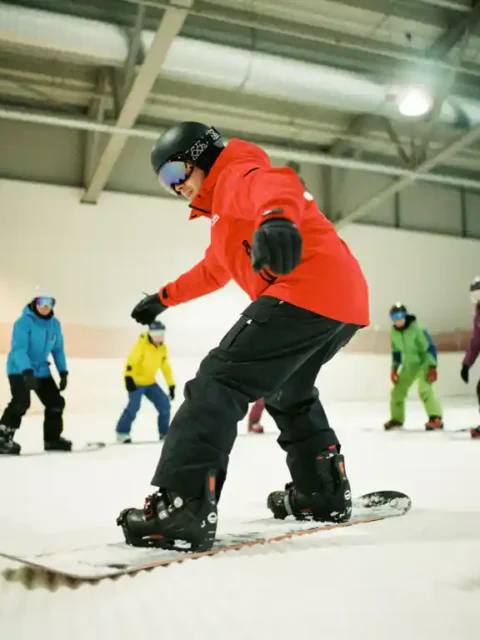 Snowboard instructeur in rode jas toont juiste houding aan beginners op kunstsneeuw in overdekte skifaciliteit