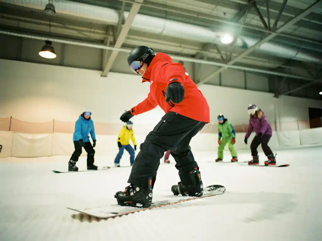 Snowboard instructeur in rode jas toont juiste houding aan beginners op kunstsneeuw in overdekte skifaciliteit