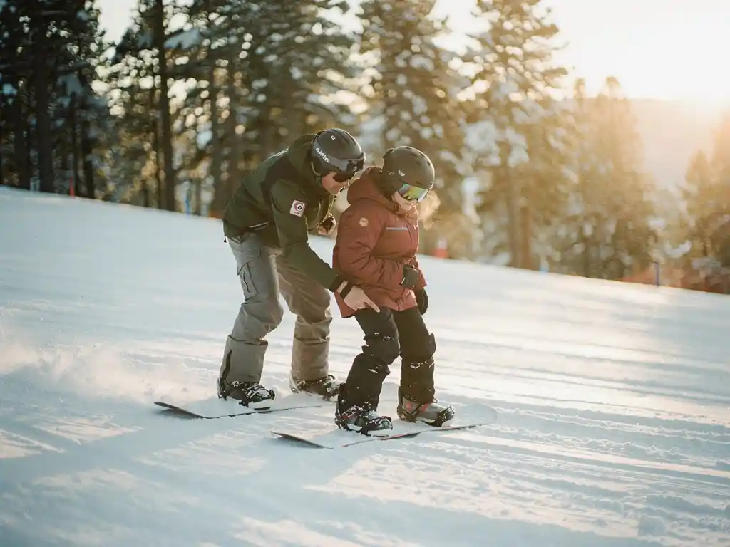 Snowboard instructeur toont juiste houding aan beginnende student op besneeuwde piste met dennenbomen op achtergrond