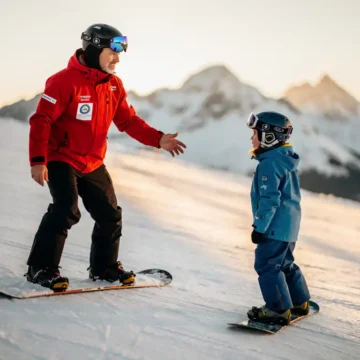 Professionele snowboard instructeur in rode jas toont juiste houding aan beginnende student op besneeuwde piste