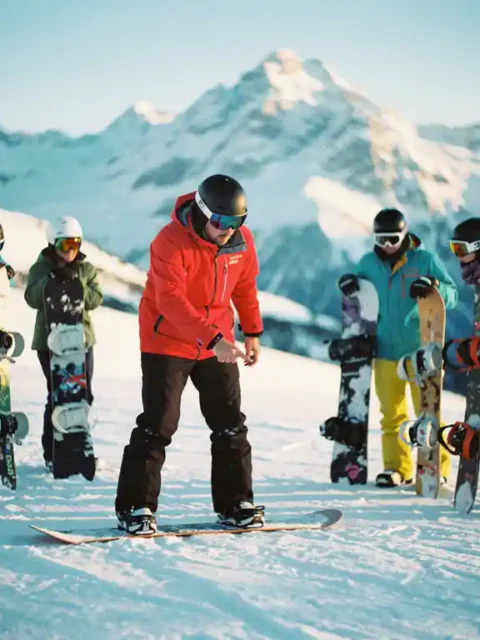 Snowboard instructeur in rode jas toont voetpositionering aan groep leerlingen op besneeuwde helling met bergen op achtergrond