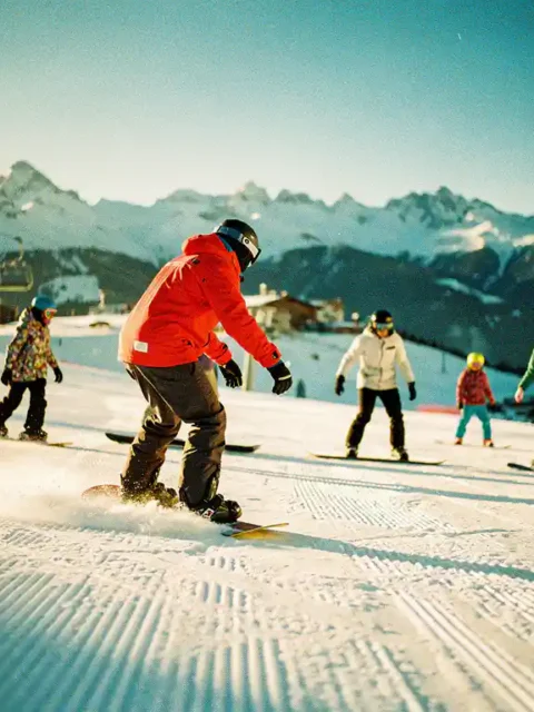 Snowboard instructeur in rode jas demonstreert techniek aan groep studenten op besneeuwde bergpiste bij zonsondergang
