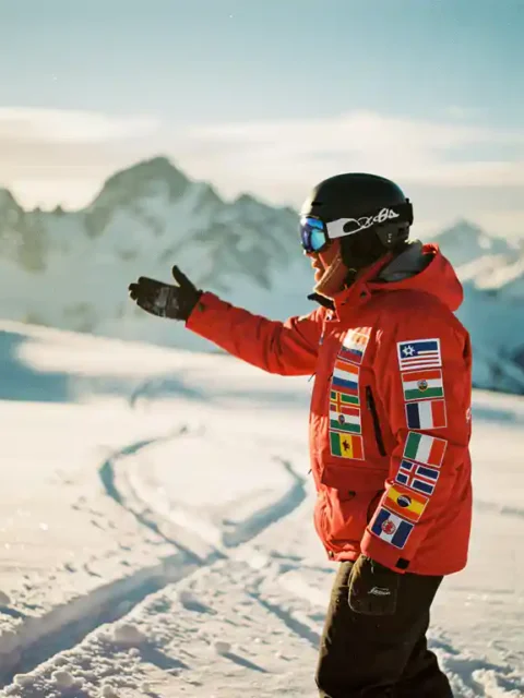 Snowboard instructeur in rode jas gebaart enthousiast op besneeuwde bergpiste met internationale vlaggen op uitrusting