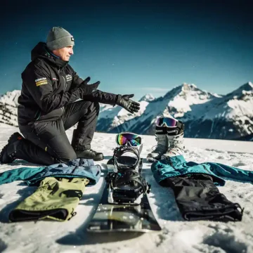 Professionele snowboard instructeur knielt naast complete snowboard uitrusting op verse poedersneeuw met bergen op achtergrond