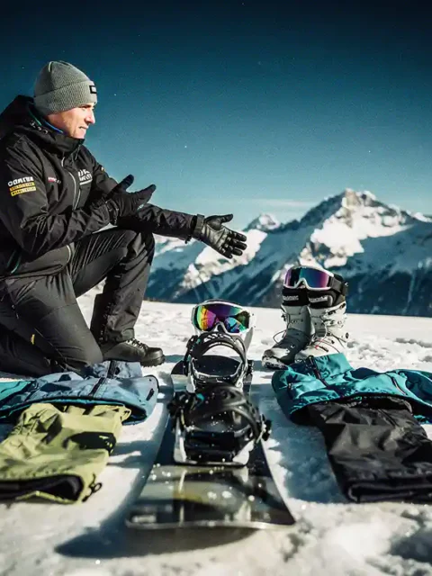 Professionele snowboard instructeur knielt naast complete snowboard uitrusting op verse poedersneeuw met bergen op achtergrond