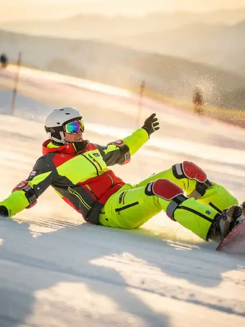 Snowboard instructor demonstrating safe falling technique on mountain slope, wearing bright safety gear and helmet.