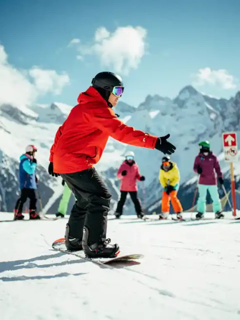 Snowboard instructor in red jacket teaching proper stance to students on snowy mountain slope with blue sky backdrop.