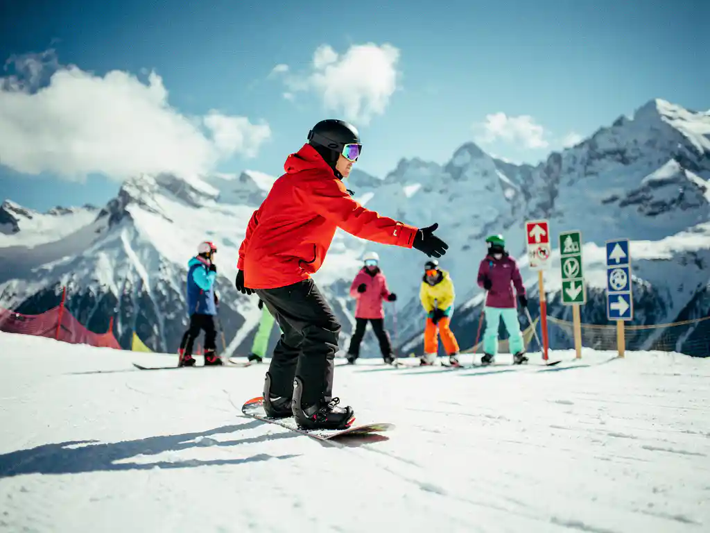 Snowboard instructor in red jacket teaching proper stance to students on snowy mountain slope with blue sky backdrop.