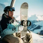 Snowboard-Lehrer demonstriert korrekte Tragetechnik im Skilift mit vertikal gehaltenen Board vor Bergpanorama