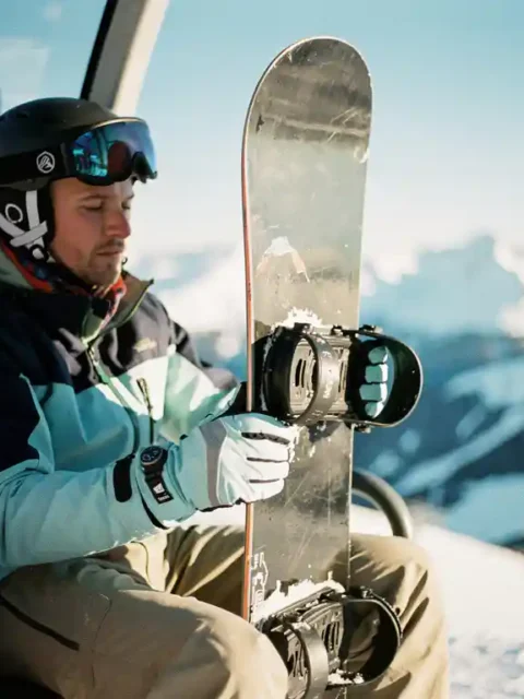 Snowboard-Lehrer demonstriert korrekte Tragetechnik im Skilift mit vertikal gehaltenen Board vor Bergpanorama