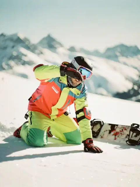 Snowboard-Instruktor demonstriert Sturztechnik auf Bergpiste, trägt Helm und Sicherheitsausrüstung, Alpen im Hintergrund