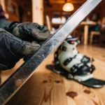 Snowboard-Metallkante wird von behandschuhten Händen in gemütlicher Skihütte inspiziert, Boots und Bindung im Hintergrund