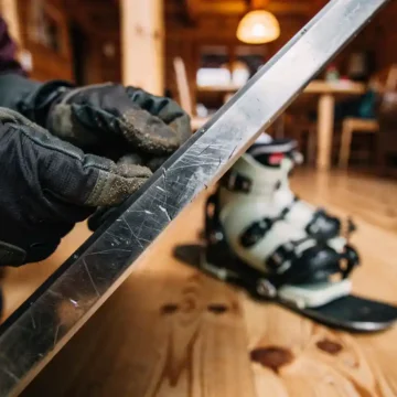 Snowboard-Metallkante wird von behandschuhten Händen in gemütlicher Skihütte inspiziert, Boots und Bindung im Hintergrund