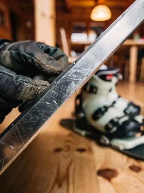 Snowboard-Metallkante wird von behandschuhten Händen in gemütlicher Skihütte inspiziert, Boots und Bindung im Hintergrund