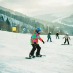 Snowboard-Lehrer zeigt korrekte Haltung auf Anfängerpiste mit grünen und blauen Markierungen im Hintergrund