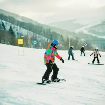 Snowboard-Lehrer zeigt korrekte Haltung auf Anfängerpiste mit grünen und blauen Markierungen im Hintergrund