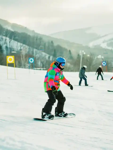 Snowboard-Lehrer zeigt korrekte Haltung auf Anfängerpiste mit grünen und blauen Markierungen im Hintergrund