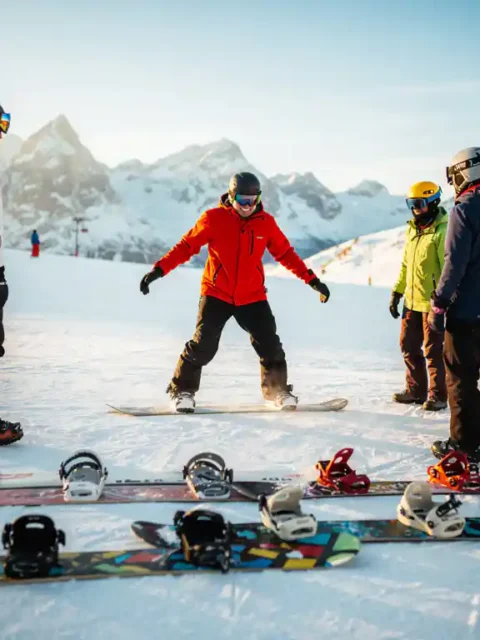 Snowboard-Lehrer in roter Jacke zeigt Anfängern die richtige Haltung auf verschneiter Piste in den Alpen
