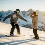 Snowboard-Lehrer mit Schutzbrille zeigt Technik, Schüler ohne Augenschutz blinzelt im Sonnenlicht auf verschneiter Piste