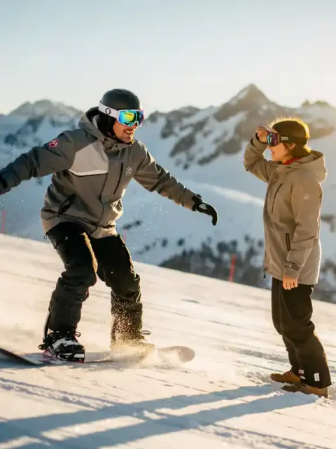 Snowboard-Lehrer mit Schutzbrille zeigt Technik, Schüler ohne Augenschutz blinzelt im Sonnenlicht auf verschneiter Piste