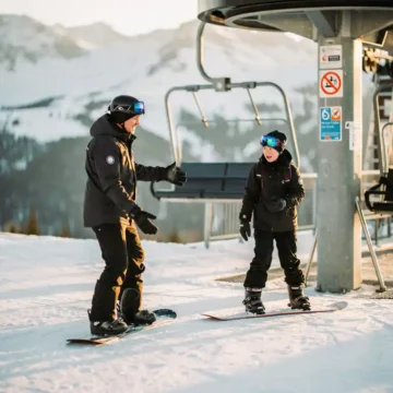Snowboard-Instruktor zeigt nervösem Anfänger das Einsteigen in den Sessellift vor verschneiten Berghängen