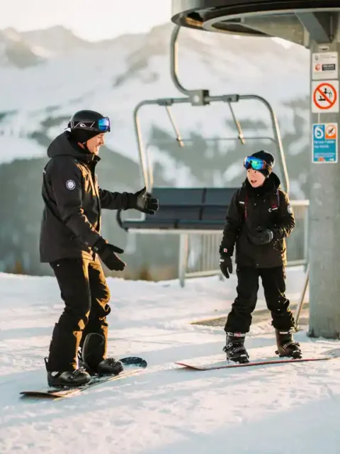 Snowboard-Instruktor zeigt nervösem Anfänger das Einsteigen in den Sessellift vor verschneiten Berghängen