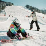 Beginnende snowboarder past bindingen aan terwijl instructeur juiste houding demonstreert op besneeuwde bergpiste