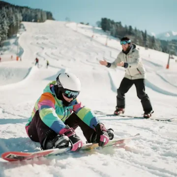 Beginnende snowboarder past bindingen aan terwijl instructeur juiste houding demonstreert op besneeuwde bergpiste