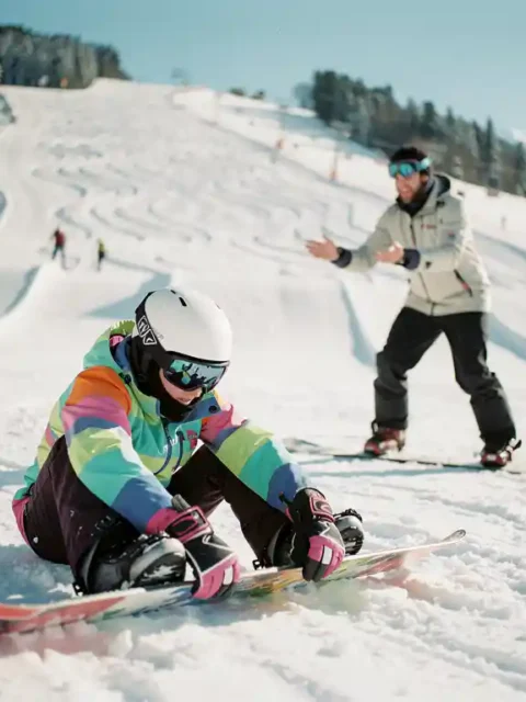 Beginnende snowboarder past bindingen aan terwijl instructeur juiste houding demonstreert op besneeuwde bergpiste