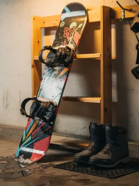 Snowboard tegen houten rek in garage met laarzen, helm en bindingen waar sneeuw vanaf smelt