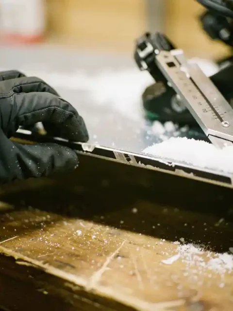 Gehandschoende handen onderzoeken versleten metalen rand van snowboard met professioneel slijpgereedschap in werkplaats