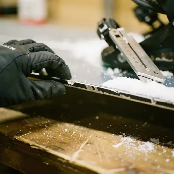 Gehandschoende handen onderzoeken versleten metalen rand van snowboard met professioneel slijpgereedschap in werkplaats