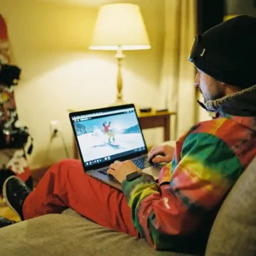 Beginnende snowboarder bekijkt tutorial op laptop in warme woonkamer, snowboard en laarzen tegen muur