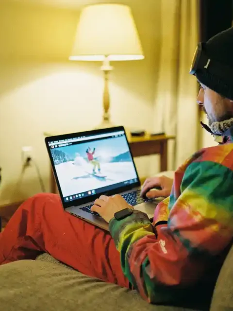 Beginnende snowboarder bekijkt tutorial op laptop in warme woonkamer, snowboard en laarzen tegen muur