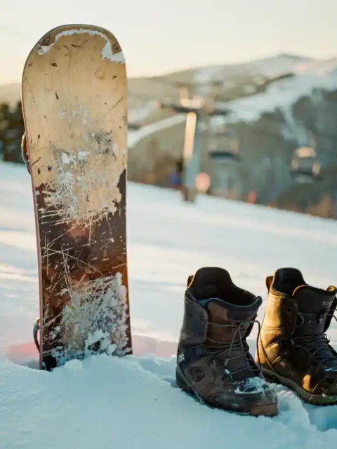 Planche de snowboard usagée appuyée contre la neige fraîche avec bottes à proximité, remontées mécaniques en arrière-plan