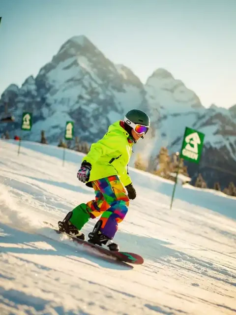 Snowboarder in felgekleurde beginneruitrusting glijdt over groene piste met besneeuwde bergen op achtergrond