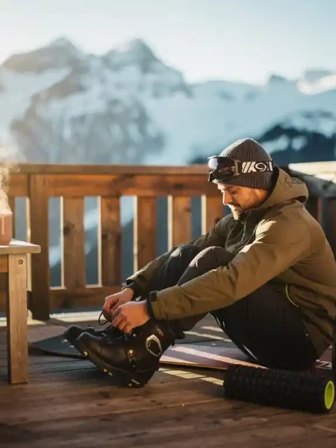 Snowboardeur se reposant sur terrasse en bois avec chocolat chaud, montagnes enneigées en arrière-plan, lumière dorée