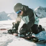 Snowboarder kneeling in snow checking binding straps and boot connections with snow-capped mountains in background.