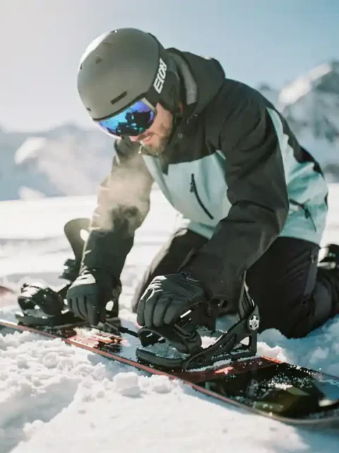 Snowboarder kneeling in snow checking binding straps and boot connections with snow-capped mountains in background.