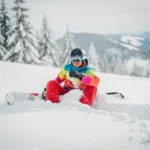 Snowboardeur en équipement coloré assis dans la poudreuse après une chute, montagnes enneigées en arrière-plan