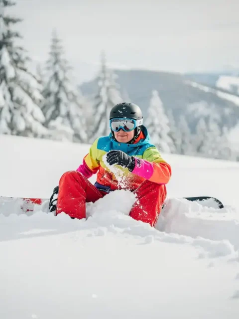 Snowboardeur en équipement coloré assis dans la poudreuse après une chute, montagnes enneigées en arrière-plan