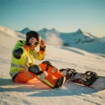 Snowboardeur en équipement de sécurité ajustant son casque sur une pente enneigée avec planche à côté, montagnes en arrière-plan