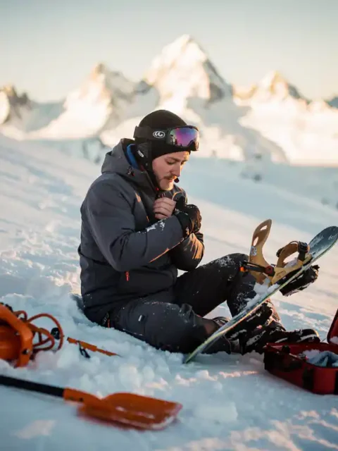 Snowboarder zit in poedersneeuw met gebroken binding en beschermend gehouden pols, reddingsuitrusting verspreid in berglandschap