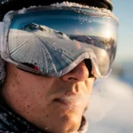Snowboarder wearing reflective goggles on snowy mountain slope with visible breath mist in cold winter sunlight