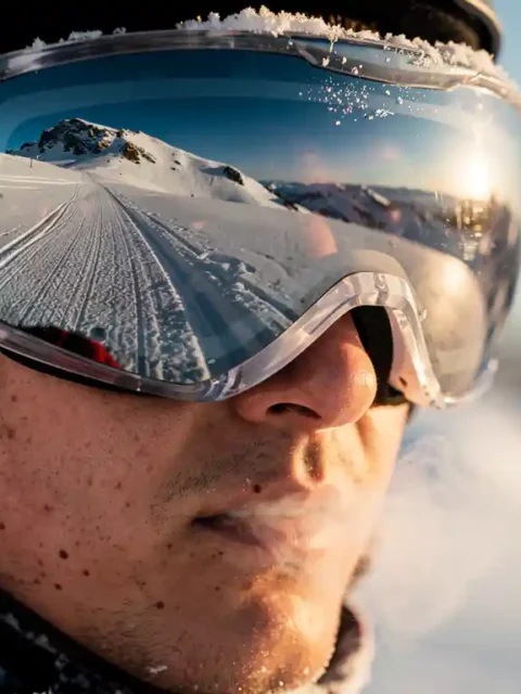 Snowboarder wearing reflective goggles on snowy mountain slope with visible breath mist in cold winter sunlight