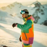 Snowboarder kijkt op horloge op besneeuwde bergpiste tijdens gouden uur, met dramatische schaduwen en bergtoppen op achtergrond