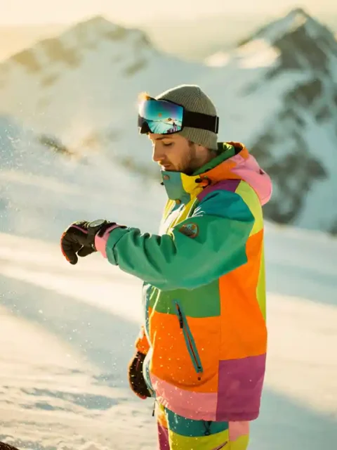 Snowboarder kijkt op horloge op besneeuwde bergpiste tijdens gouden uur, met dramatische schaduwen en bergtoppen op achtergrond