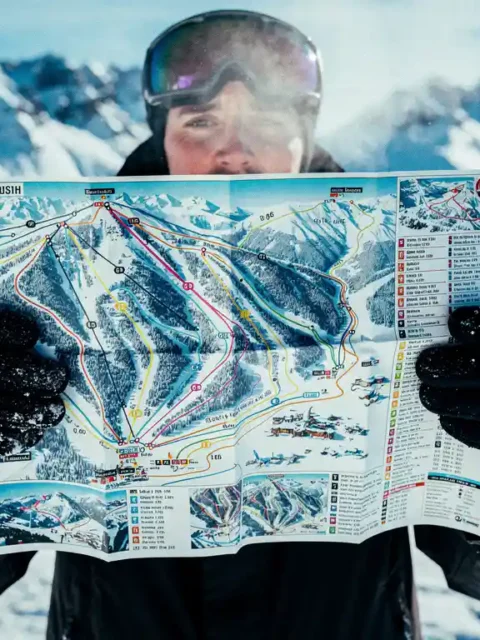 Snowboarder in goggles holding colorful piste map with trail markings against snowy mountain backdrop on sunny day