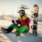 Snowboarder in colorful winter gear reading insurance document on snowy mountain slope with ski lift in background