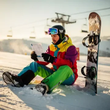 Snowboarder in colorful winter gear reading insurance document on snowy mountain slope with ski lift in background